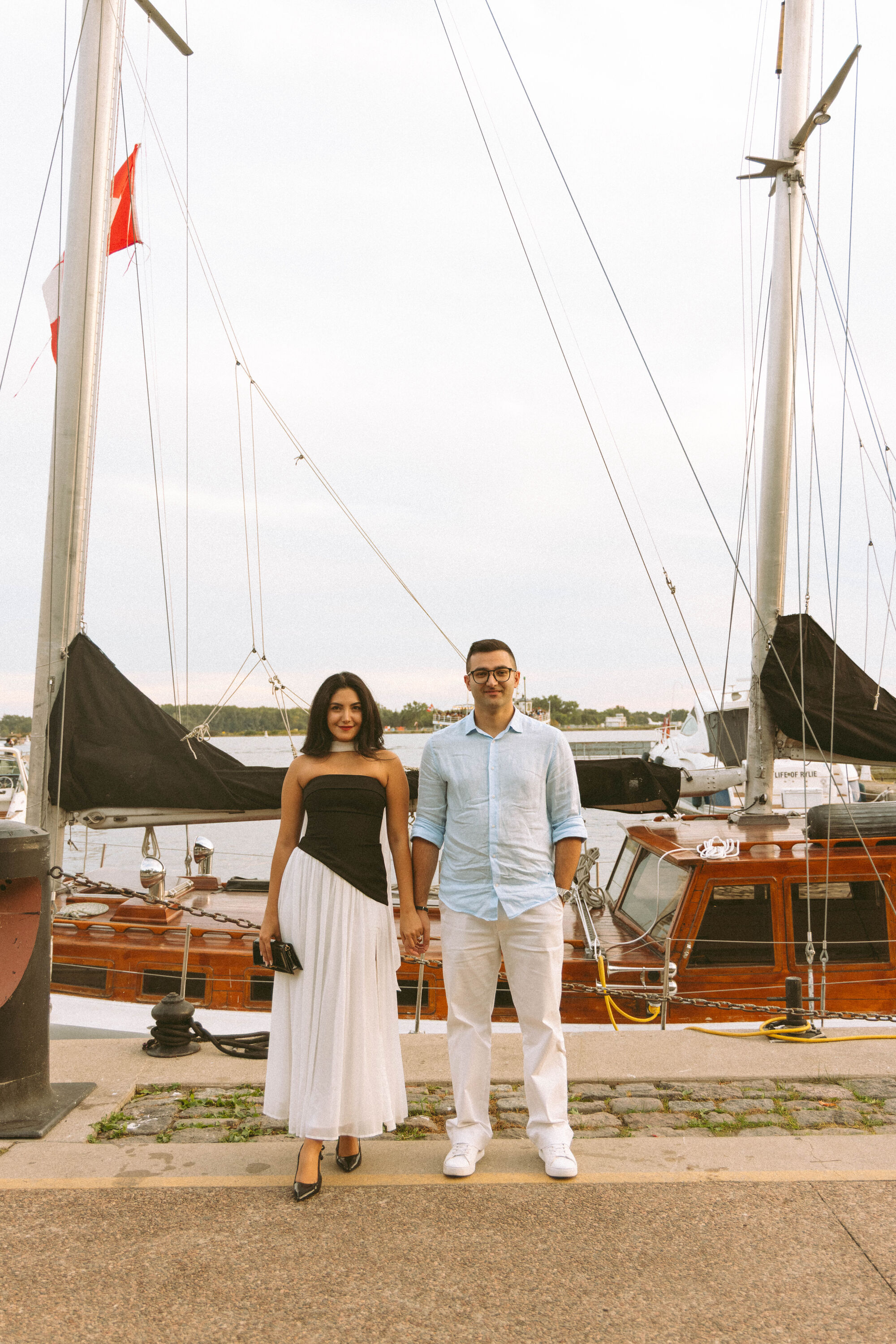 Majid and Mehrnaz standing side by side beside a wooden sailboat in the harbour